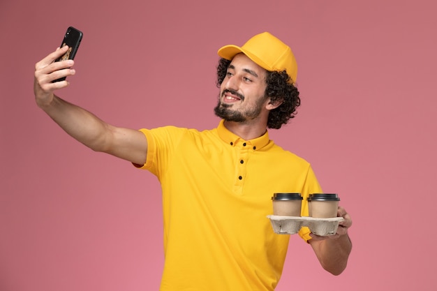 Vue avant du courrier masculin en uniforme jaune tenant des tasses de café de livraison marron prenant photo sur mur rose