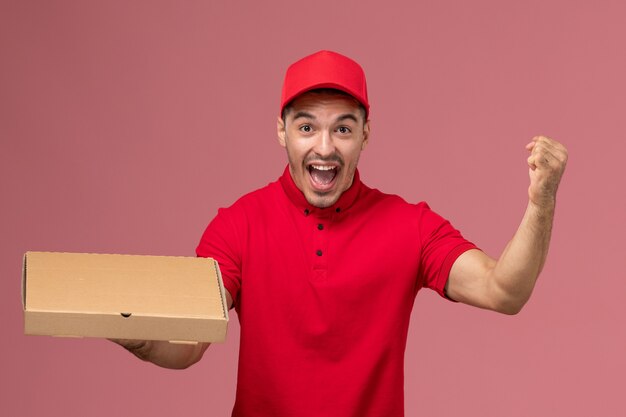 Vue avant du courrier masculin en uniforme rouge et cape se réjouissant et tenant la boîte de livraison de nourriture sur l'emploi de travailleur de mur rose