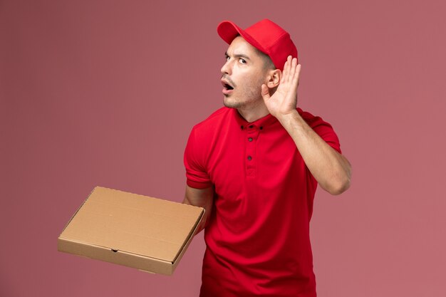 Vue avant du courrier masculin en uniforme rouge et cape tenant la boîte de livraison de nourriture essayant d'entendre sur l'emploi de travailleur de mur rose