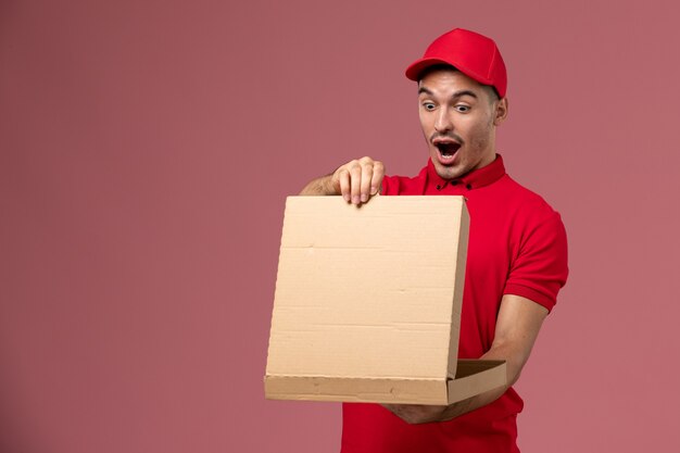 Vue avant du courrier masculin en uniforme rouge et cape tenant la boîte de nourriture l'ouvrant sur le travail de travailleur mural rose clair