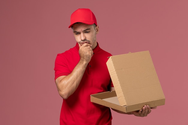 Vue avant du courrier masculin en uniforme rouge et cape tenant la boîte de nourriture et de la pensée sur le travail de service de mur rose uniforme de livraison masculine