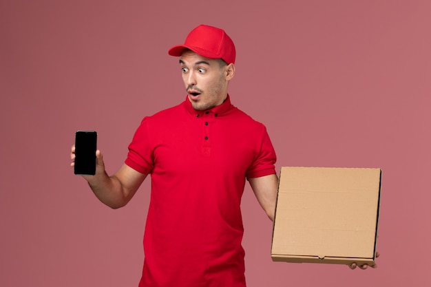 Vue avant du courrier masculin en uniforme rouge et cape tenant la boîte de nourriture avec téléphone sur le mur rose emploi de service de l'homme uniforme de livraison