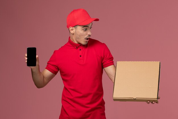 Vue avant du courrier masculin en uniforme rouge et cape tenant la boîte de nourriture avec téléphone sur le plancher rose de travail de service de l'homme uniforme de livraison
