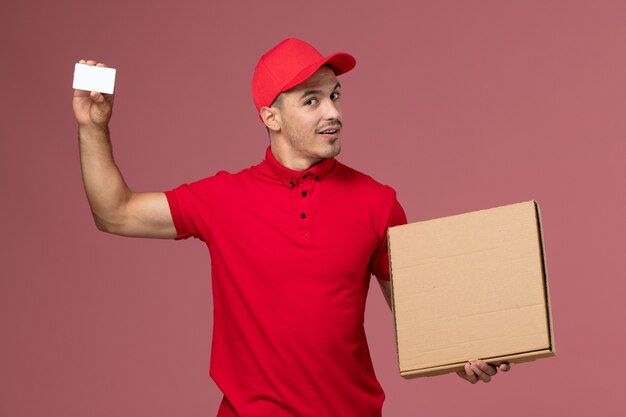 Vue avant du courrier masculin en uniforme rouge et cape tenant une carte blanche et une boîte de nourriture sur un mur rose