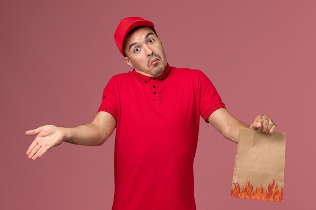 Vue avant du courrier masculin en uniforme rouge et cape tenant le paquet alimentaire papier posant sur le mur rose