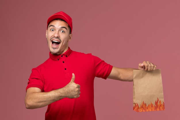 Vue avant du courrier masculin en uniforme rouge et cape tenant le paquet alimentaire se réjouissant sur le mur rose