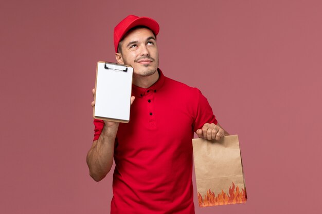 Vue avant du courrier masculin en uniforme rouge et cape tenant le paquet de nourriture et le bloc-notes pensant sur le mur rose