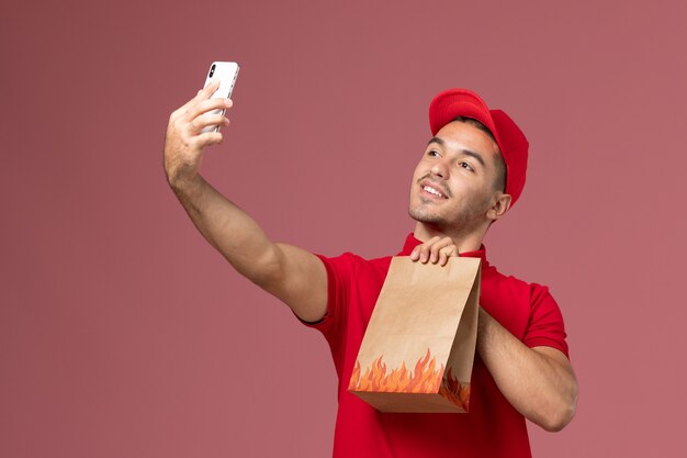 Vue avant du courrier masculin en uniforme rouge et cape tenant le paquet de nourriture et prenant selfie avec elle sur le mur rose