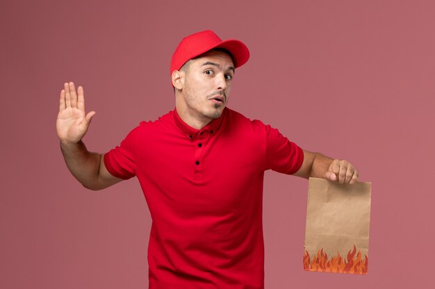 Vue avant du courrier masculin en uniforme rouge et cape tenant un paquet de papier alimentaire sur le mur rose