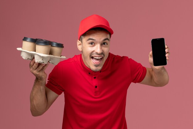 Vue avant du courrier masculin en uniforme rouge et cape tenant des tasses de café de livraison avec téléphone sur le mur rose