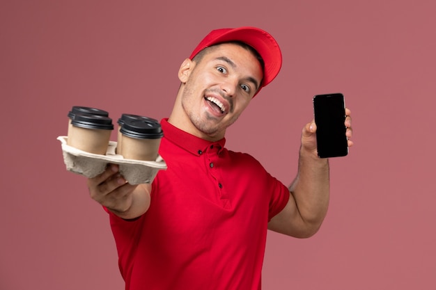 Vue avant du courrier masculin en uniforme rouge et cape tenant des tasses de café de livraison avec téléphone sur le mur rose