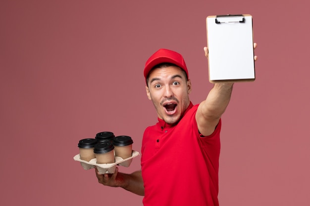 Vue avant du courrier masculin en uniforme rouge se réjouissant et tenant des tasses de café de livraison avec bloc-notes sur mur rose