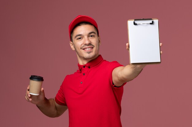 Vue avant du courrier masculin en uniforme rouge tenant la tasse de café de livraison et le bloc-notes souriant sur le travail masculin de travailleur de mur rose