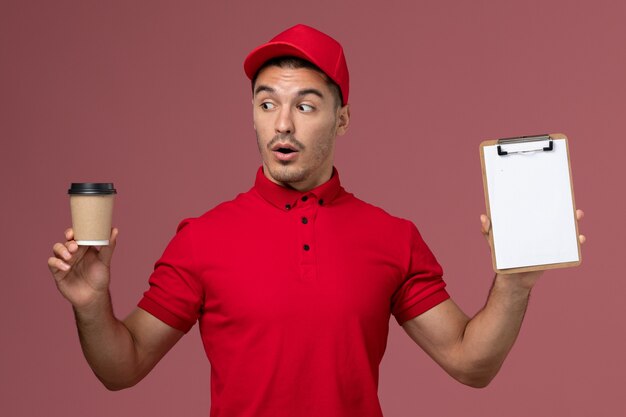 Vue avant du courrier masculin en uniforme rouge tenant la tasse de café de livraison et le bloc-notes sur le travail de travailleur de mur rose