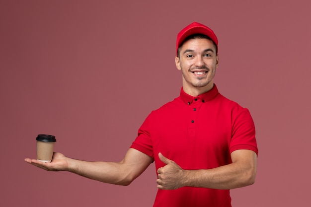 Vue avant du courrier masculin en uniforme rouge tenant la tasse de café de livraison sur le mur rose travailleur de l'emploi