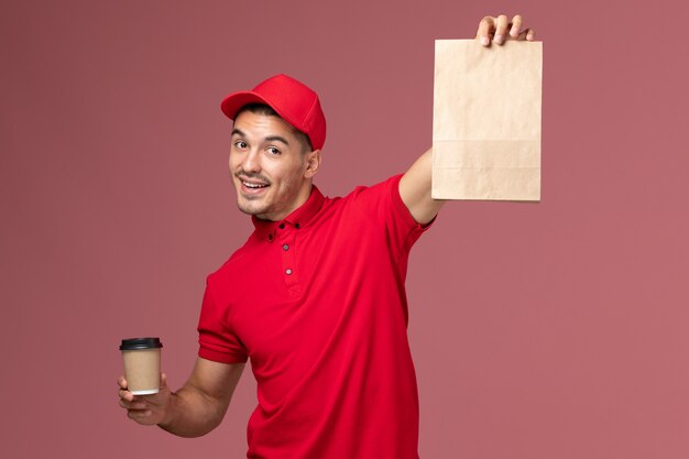 Vue avant du courrier masculin en uniforme rouge tenant la tasse de café de livraison et le paquet de nourriture sur le mur rose travailleur de livraison de service emploi uniforme