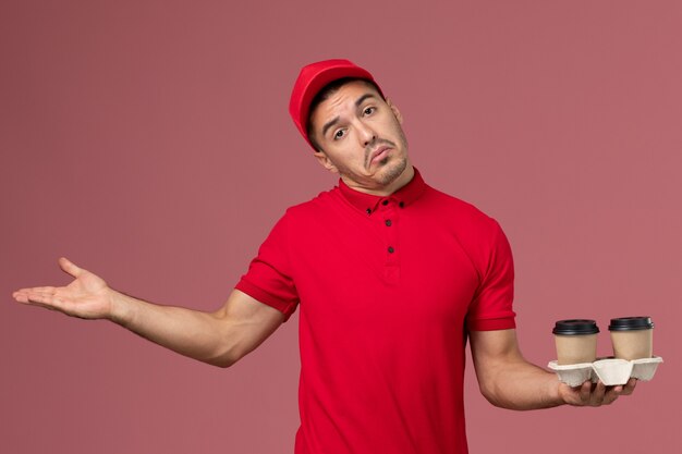 Vue avant du courrier masculin en uniforme rouge tenant des tasses de café de livraison marron sur le bureau rose mâle