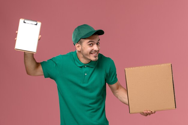 Vue avant du courrier masculin en uniforme vert tenant la boîte de nourriture avec le bloc-notes sur le fond rose clair