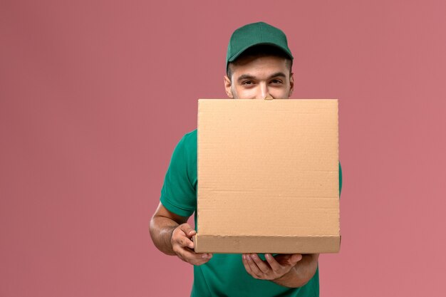 Vue avant du courrier masculin en uniforme vert tenant la boîte de nourriture et l'ouvrir sur le bureau rose clair