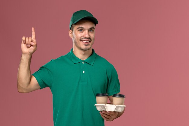 Vue avant du courrier masculin en uniforme vert tenant des tasses de café de livraison marron sur un sol rose clair