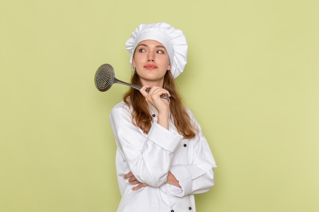 Vue avant du cuisinier femme portant un costume de cuisinier blanc tenant une grande cuillère en argent sur un mur vert clair