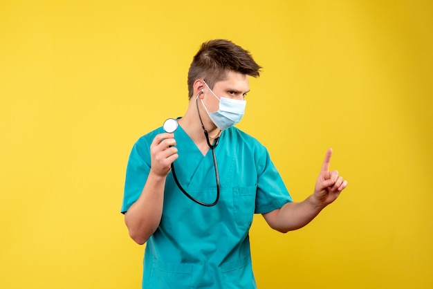 Vue avant du médecin de sexe masculin en costume médical et masque avec stéthoscope sur mur jaune