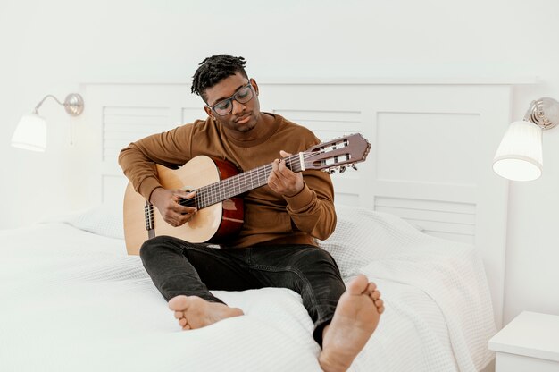 Vue avant du musicien masculin à la maison à jouer de la guitare sur le lit