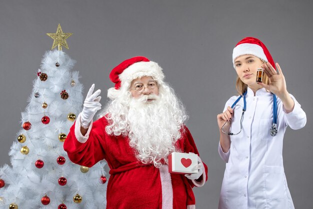 Vue avant du père Noël avec femme médecin qui tient petit flacon avec des pilules sur mur gris