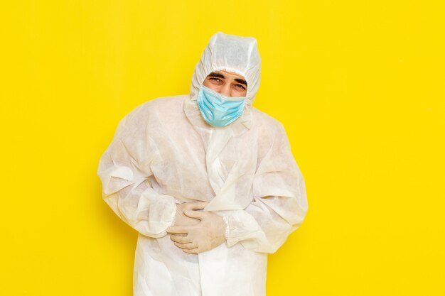 Vue avant du travailleur scientifique masculin en combinaison blanche de protection spéciale et avec masque ayant mal au ventre sur un mur jaune clair
