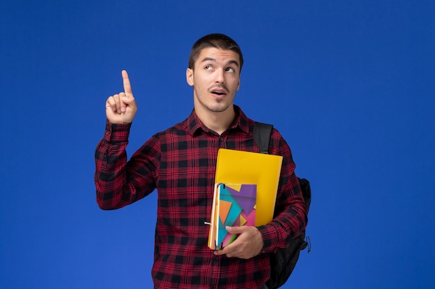 Vue avant de l'étudiant masculin en chemise à carreaux rouge avec sac à dos contenant des fichiers et des cahiers sur mur bleu clair