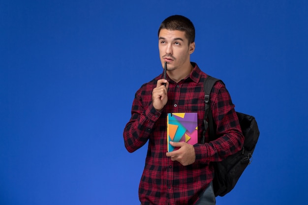 Vue avant de l'étudiant masculin en chemise à carreaux rouge avec sac à dos tenant un cahier pensant sur le mur bleu clair