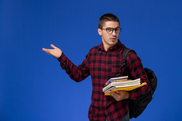 Vue avant de l'étudiant masculin en chemise à carreaux rouge avec sac à dos tenant des cahiers sur le mur bleu clair