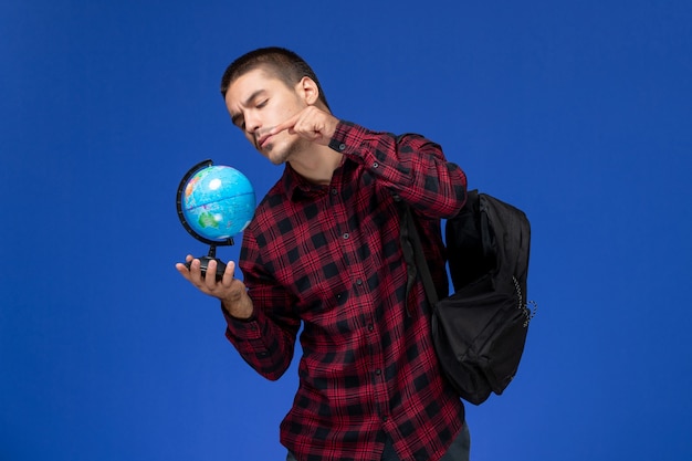 Vue avant de l'étudiant masculin en chemise à carreaux rouge avec sac à dos tenant petit globe sur mur bleu clair