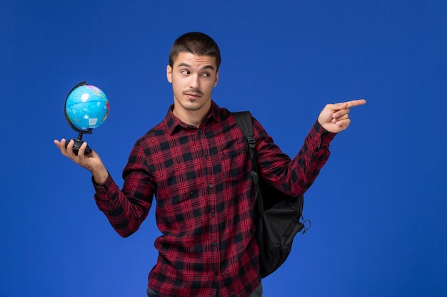 Vue avant de l'étudiant masculin en chemise à carreaux rouge avec sac à dos tenant petit globe sur mur bleu clair