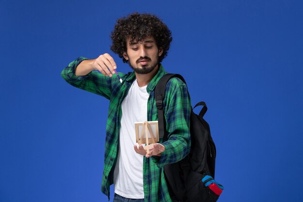 Vue avant de l'étudiant masculin en chemise à carreaux vert avec sac à dos noir tenant pompon et chevalet sur mur bleu