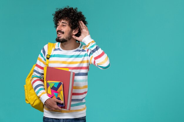 Vue avant de l'étudiant masculin en chemise rayée de couleur portant un sac à dos jaune tenant des fichiers et des cahiers essayant d'entendre sur le mur bleu