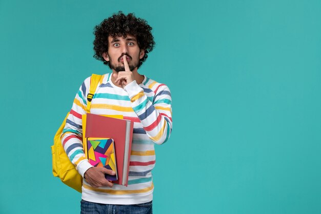 Vue avant de l'étudiant masculin en chemise rayée de couleur portant sac à dos jaune tenant des fichiers et des cahiers sur mur bleu