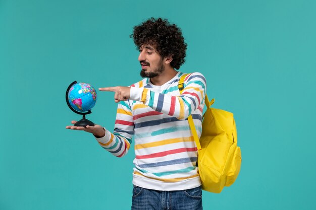 Vue avant de l'étudiant masculin en chemise rayée portant sac à dos jaune tenant petit globe rond sur le mur bleu