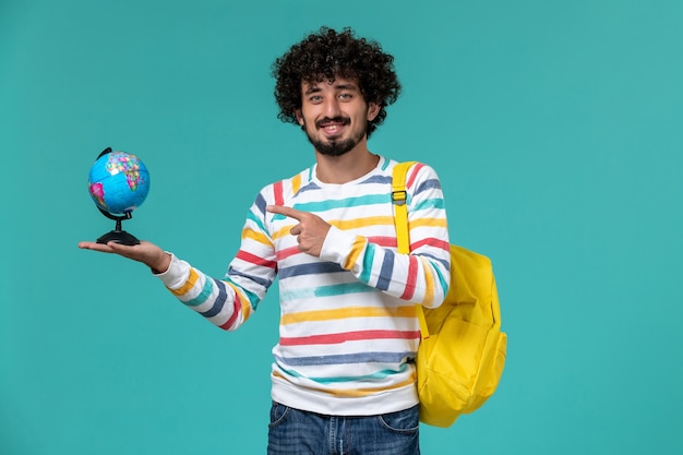 Vue avant de l'étudiant masculin en chemise rayée portant sac à dos jaune tenant petit globe rond sur le mur bleu