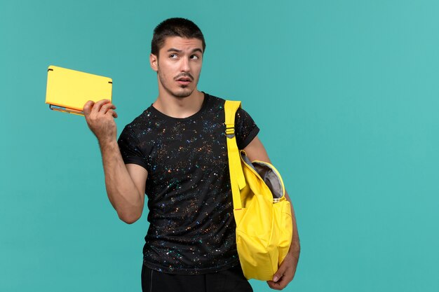 Vue avant de l'étudiant masculin en t-shirt foncé sac à dos jaune tenant un cahier sur un mur bleu clair