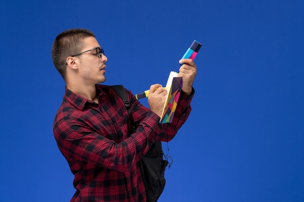 Vue avant de l'étudiant de sexe masculin en chemise à carreaux rouge avec sac à dos tenant cahier de feutres sur mur bleu clair