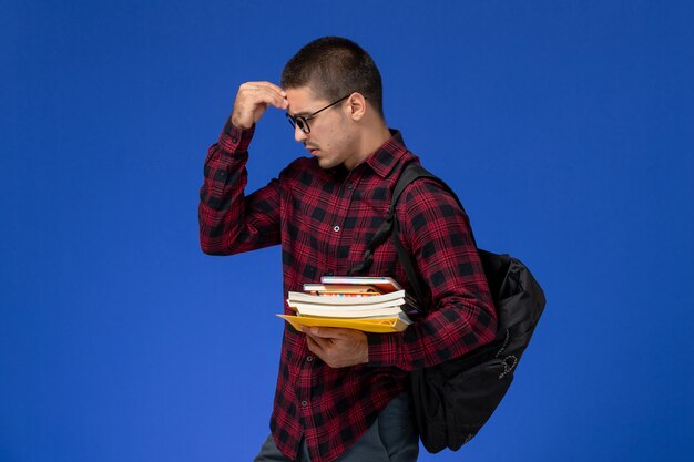 Vue avant de l'étudiant de sexe masculin en chemise à carreaux rouge avec sac à dos tenant des cahiers et des cahiers sur mur bleu clair