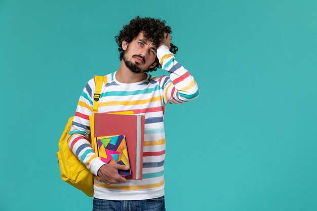 Vue avant de l'étudiant de sexe masculin en chemise rayée de couleur portant un sac à dos jaune tenant des fichiers et des cahiers stressés sur mur bleu