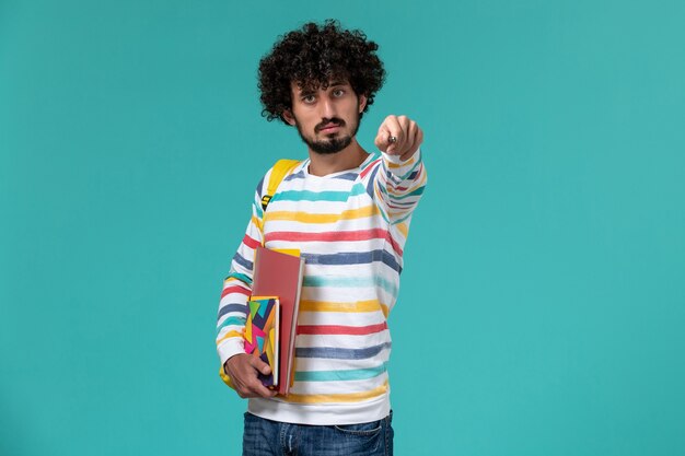 Vue avant de l'étudiant de sexe masculin portant un sac à dos jaune tenant un stylo et un cahier sur le mur bleu