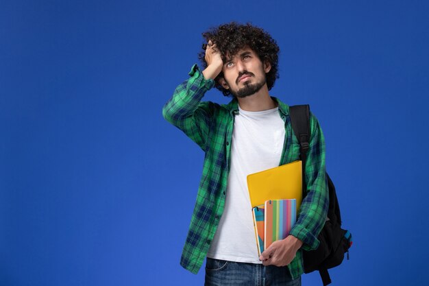 Vue avant de l'étudiant de sexe masculin portant un sac à dos noir tenant des cahiers et des fichiers ayant des maux de tête sur le mur bleu