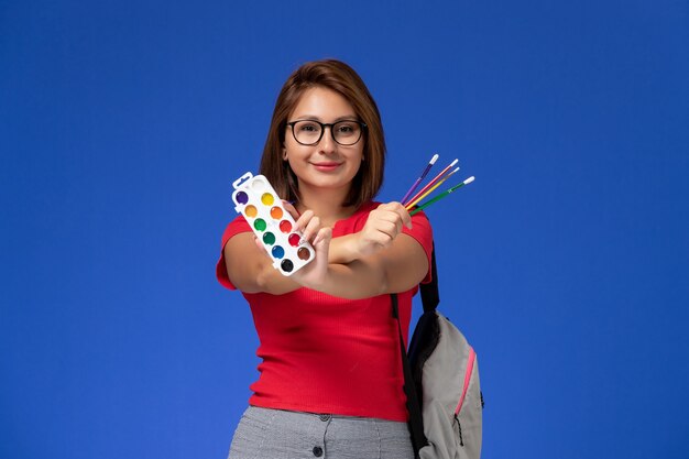 Vue avant de l'étudiante en chemise rouge avec sac à dos tenant des peintures pour le dessin et des glands sur le mur bleu