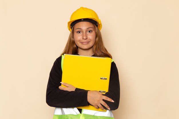 Vue avant femme constructeur en casque jaune tenant un document jaune avec sourire sur mur blanc