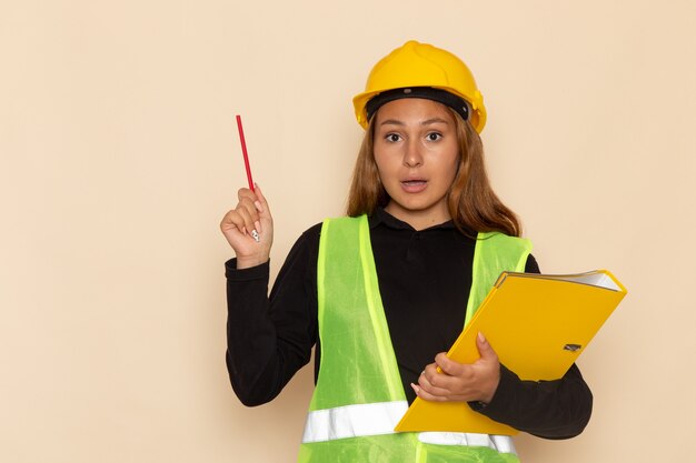 Vue avant femme constructeur en casque jaune tenant fichier jaune et crayon sur mur blanc