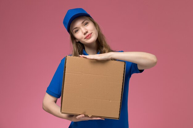 Vue avant femme courrier en uniforme bleu tenant la boîte de nourriture avec le sourire sur le bureau rose entreprise uniforme de service d'emploi