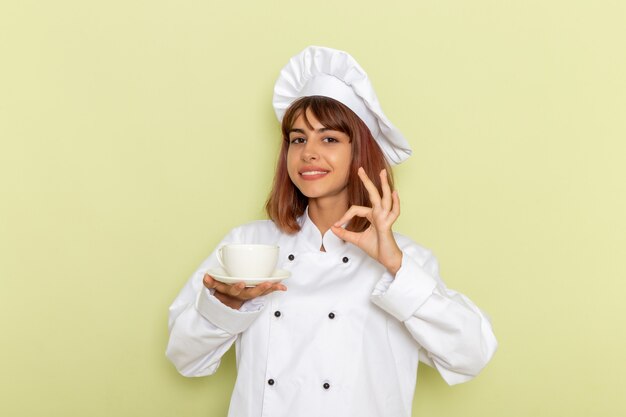 Vue avant femme cuisinier en costume de cuisinier blanc, boire du thé sur un bureau vert